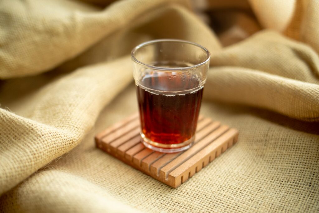 A clear glass filled with warm tea resting on a wooden coaster and burlap fabric.