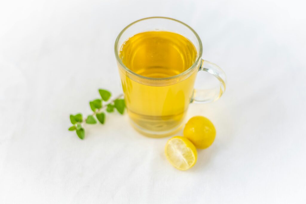 A clear glass of lemon herbal tea accompanied by fresh herbs on a white surface, offering a refreshing drink.