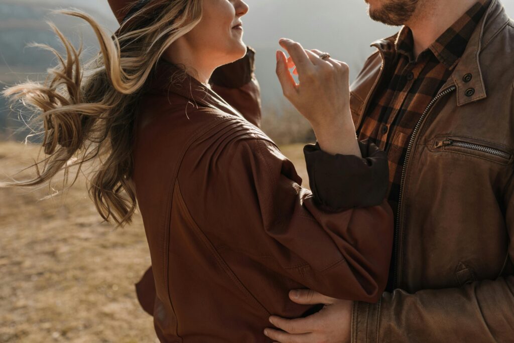 A man and woman embrace in cozy leather jackets on a windy autumn day outdoors, enjoying each other's company.